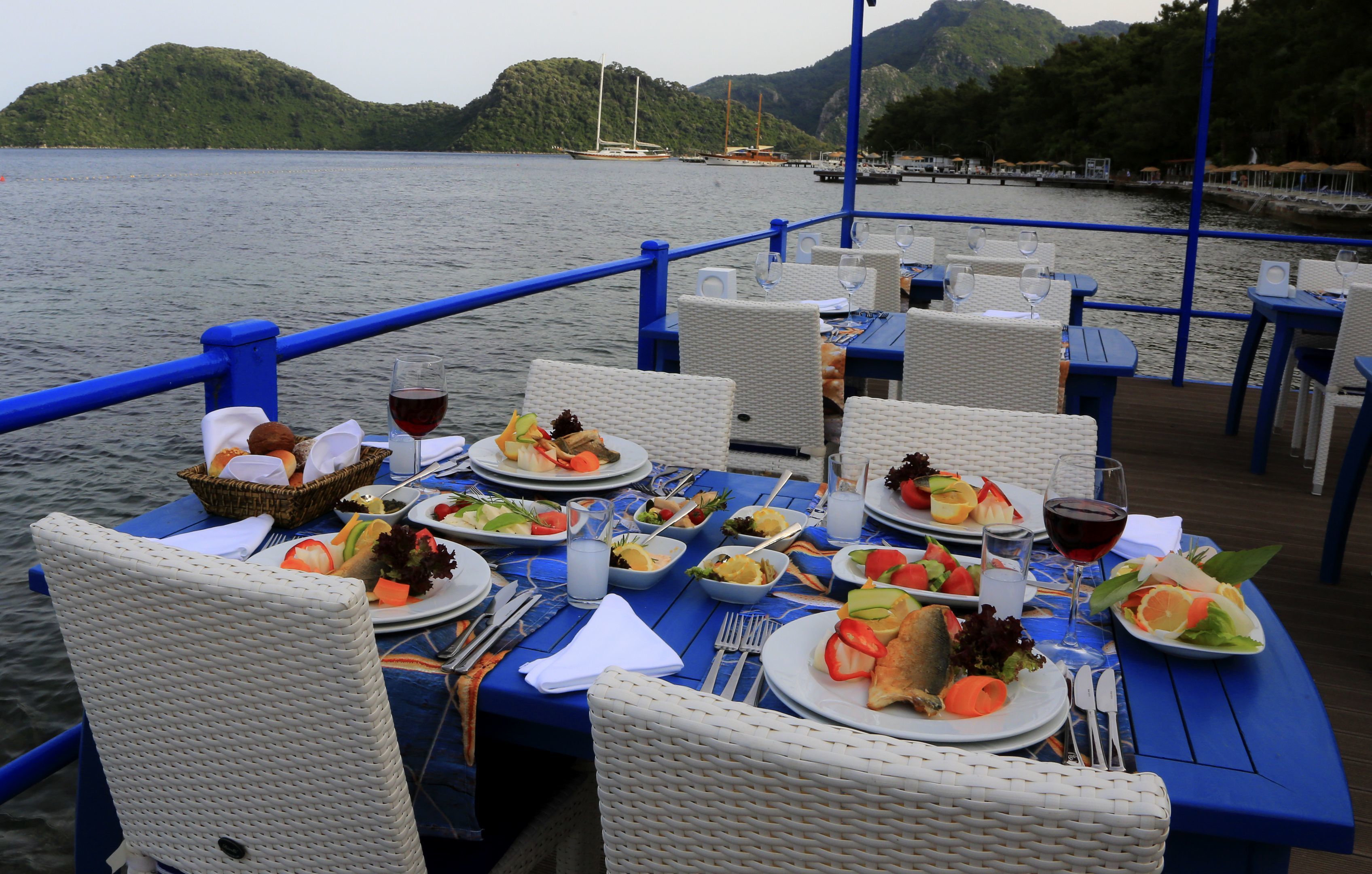 A dining table with multiple plates of food, glasses of wine, and wicker chairs on a boat deck with a view of the water and hills.
