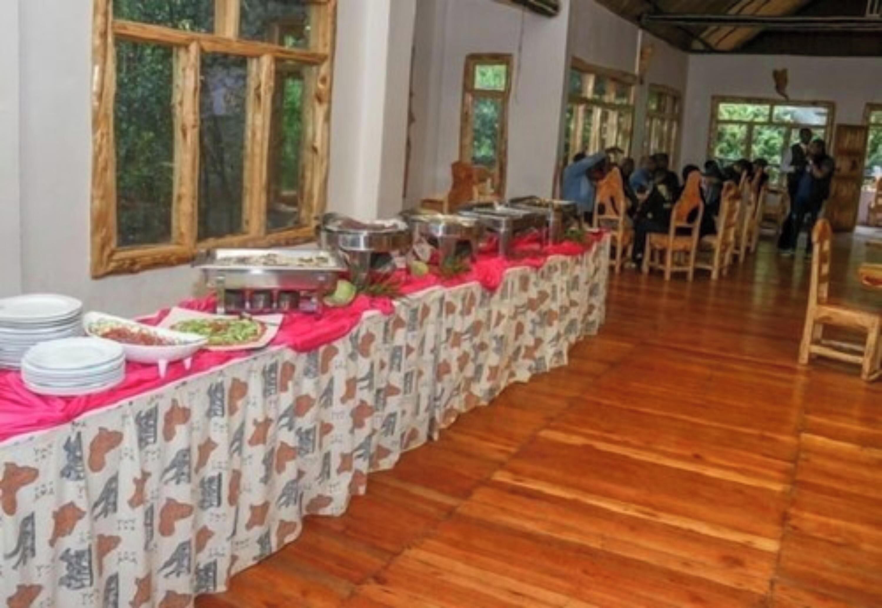 A long table with food containers and plates, set in a room with wooden floors and large windows.