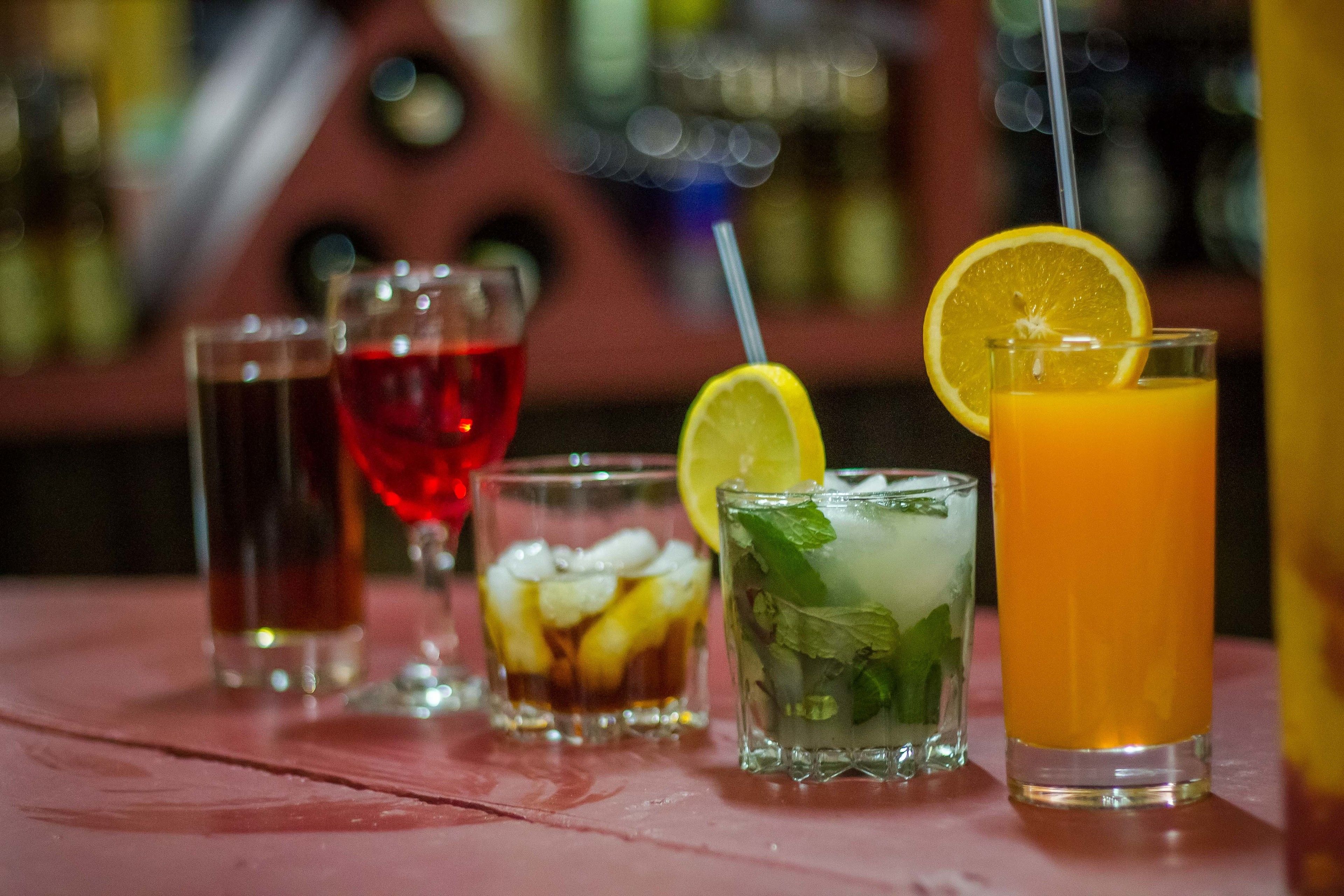 Um bar com diversos bebidas, incluindo um cocktail vermelho, um copo com gelo e limão, e um copo alto de sumo de laranja.