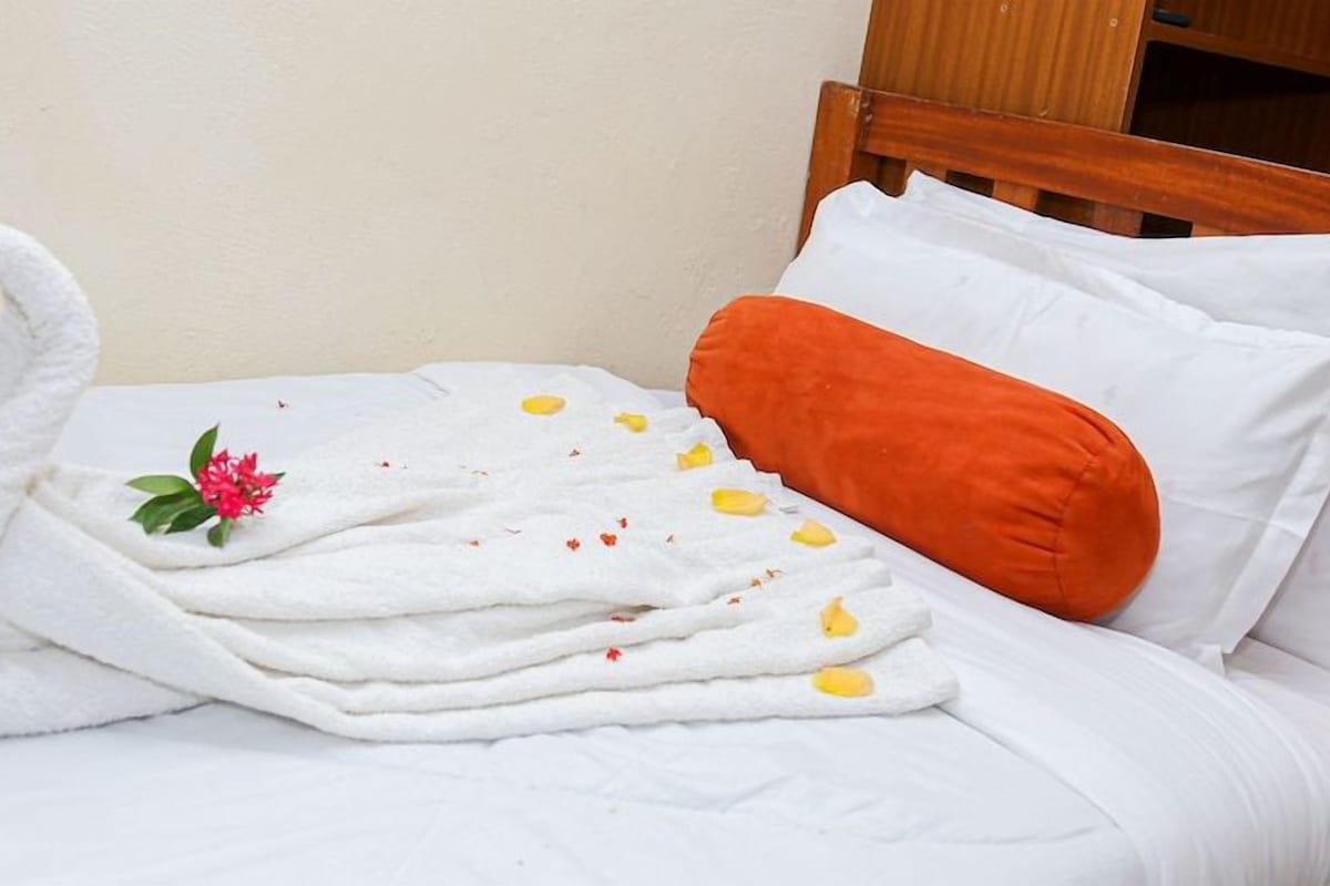 A neatly made bed with white linens, a decorative swan towel arrangement, a single pink flower, and an orange pillow.
