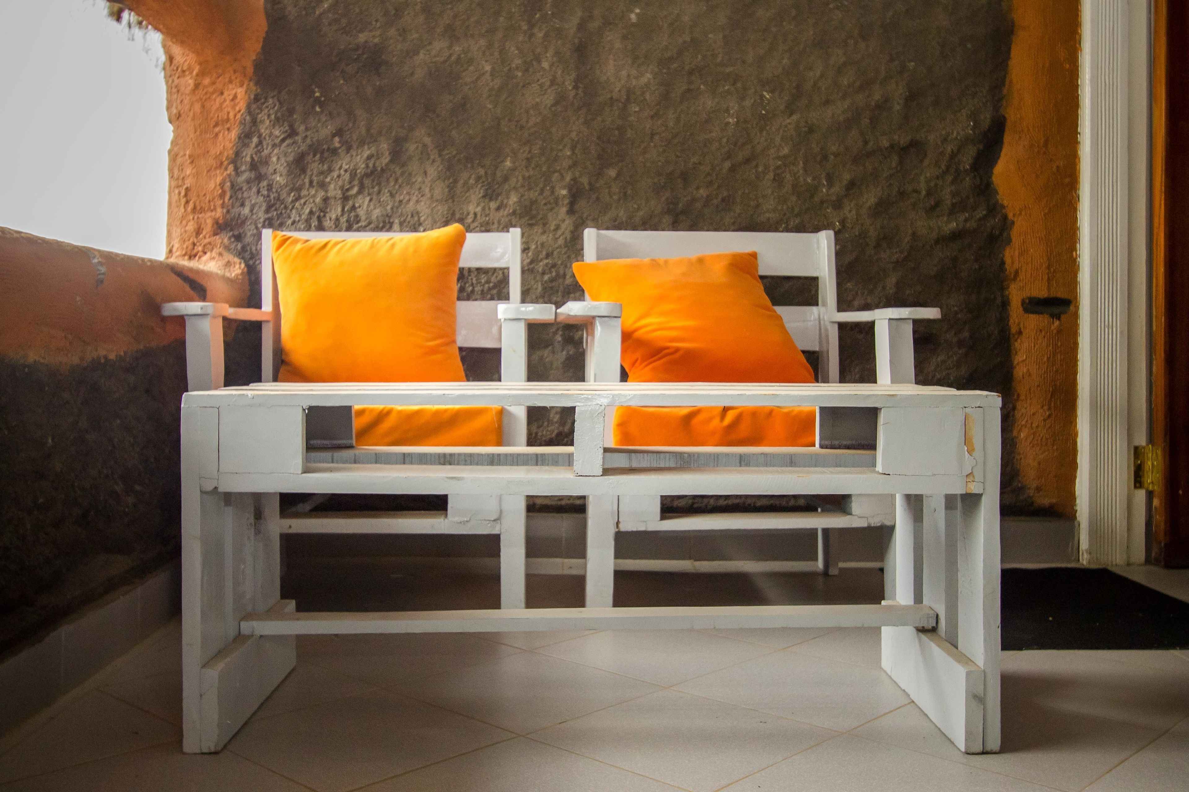 A white wooden bed with two orange pillows against a textured wall.