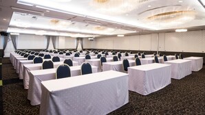 Une salle de conférence avec des rangées de tables et de chaises, un écran de projection et un plafond orné de lustres.