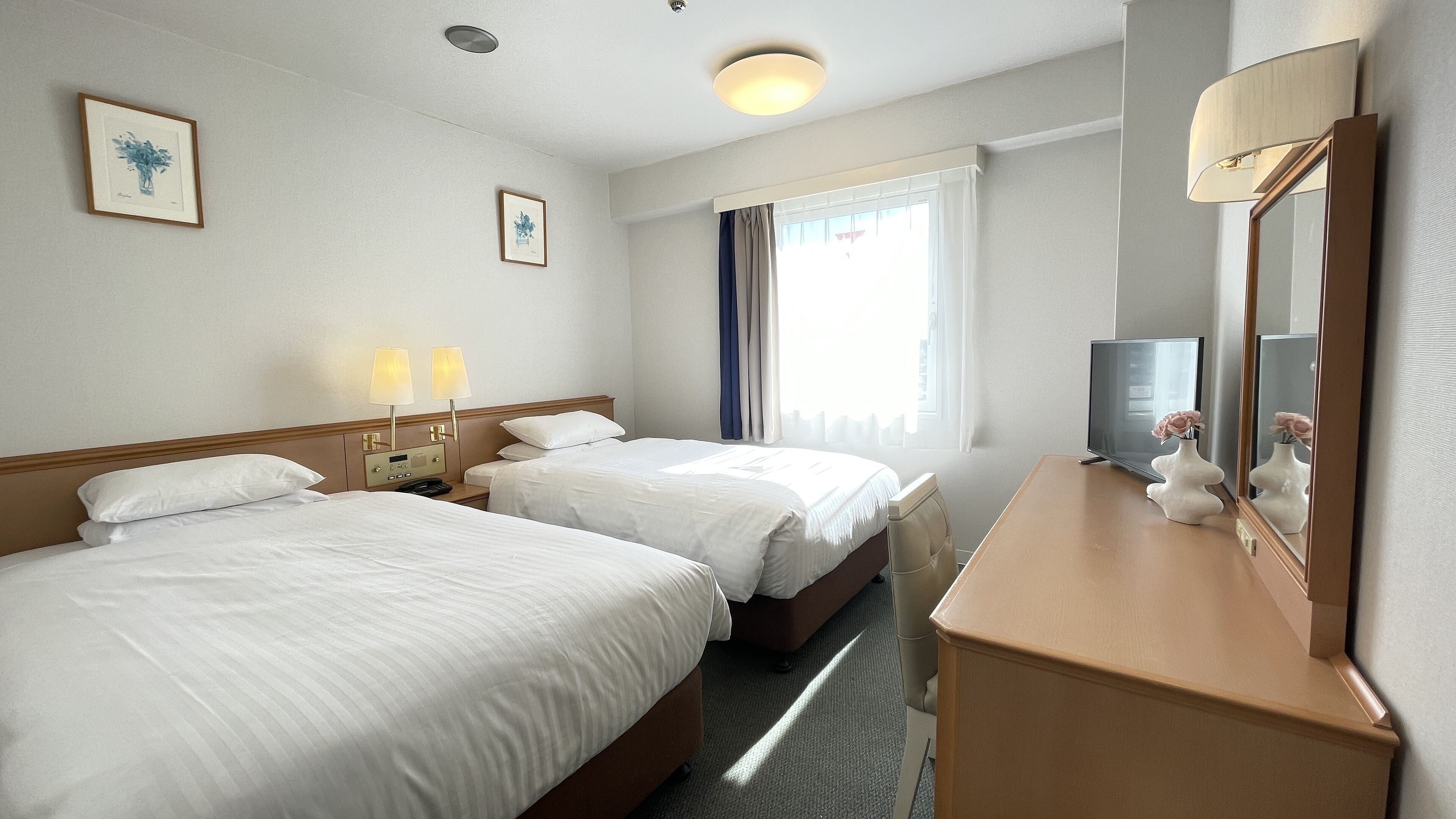 A hotel room with two beds, a bedside table with lamps, a TV, a mirror, and artwork on the walls.