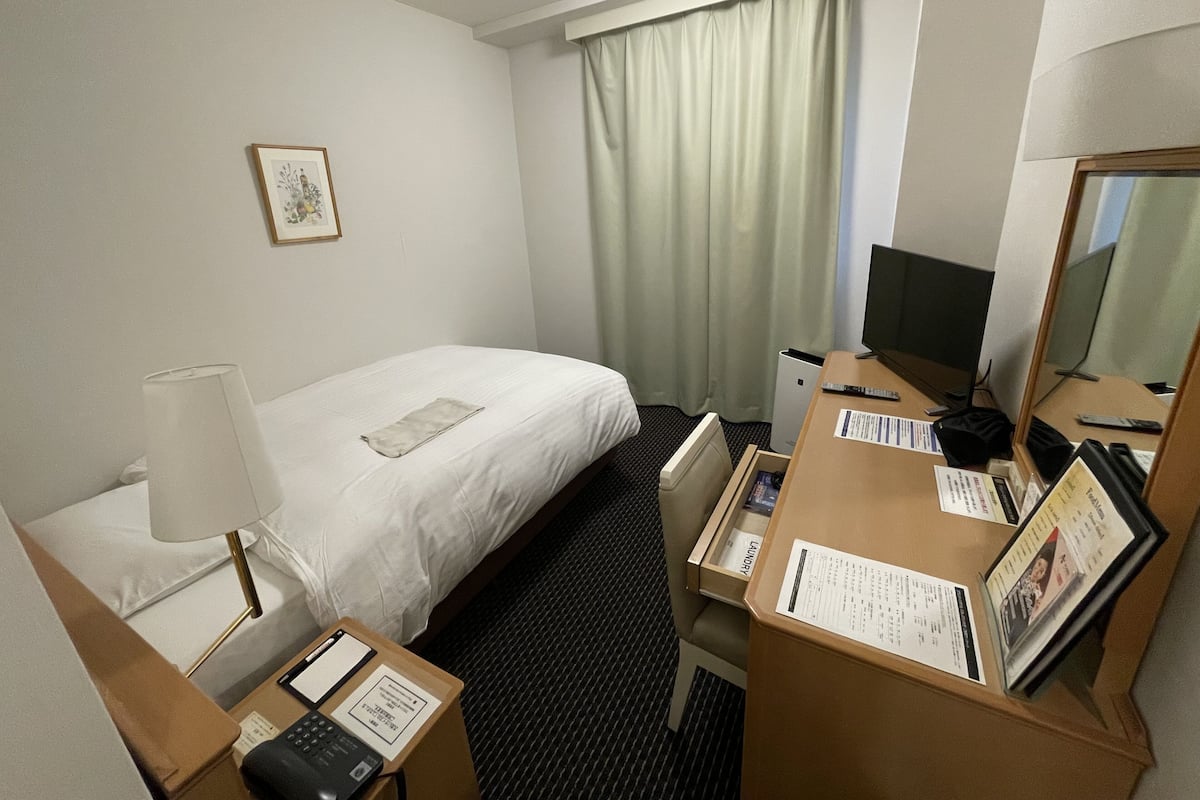 A hotel room with a bed, a desk with a computer, a telephone, a lamp, and a window with curtains.
