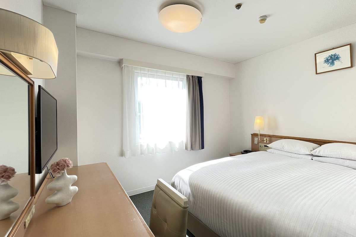A hotel room with a bed, a desk, a mirror, a lamp, and a window with curtains.