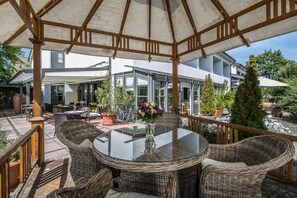 A covered outdoor patio with wicker furniture and a glass table, surrounded by potted plants and a view of a modern house. - Hotel am Krone Park (Olching)