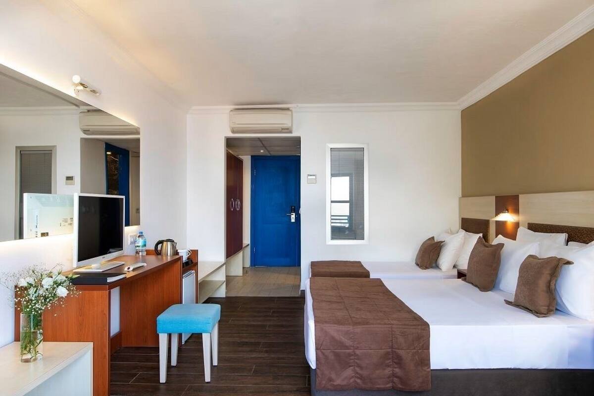 A hotel room with a bed, a desk, a chair, a small table with a vase of flowers, and a blue door in the background.