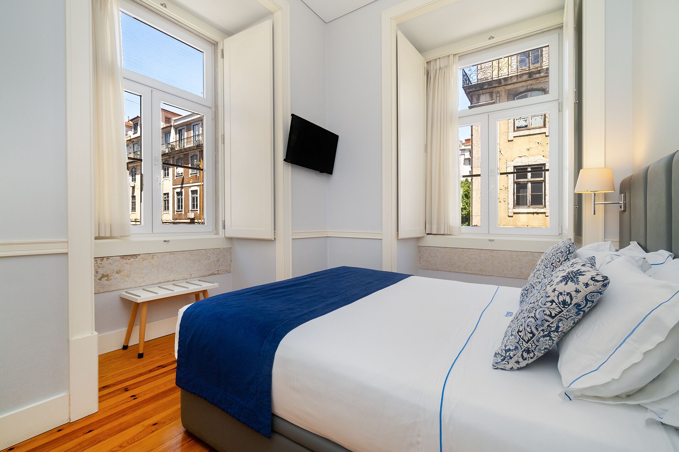 A bedroom with a bed, a TV mounted on the wall, and a window with a view of buildings.