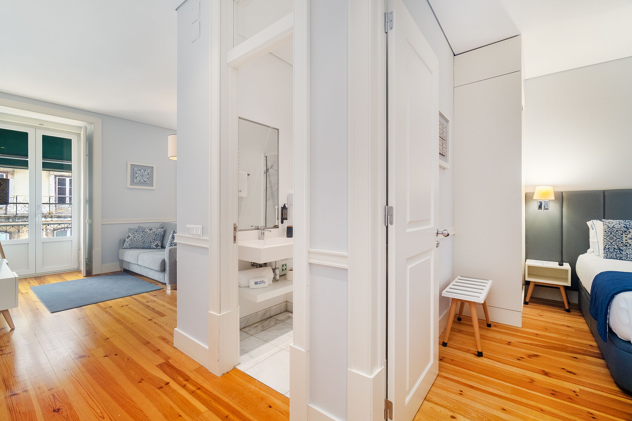 A modern bedroom with a bed, bedside table, and a small stool. There is a bathroom with a sink and mirror. The room has wooden flooring and white walls.