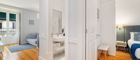 A modern bedroom with a bed, bedside table, and a small stool. There is a bathroom with a sink and mirror. The room has wooden flooring and white walls.