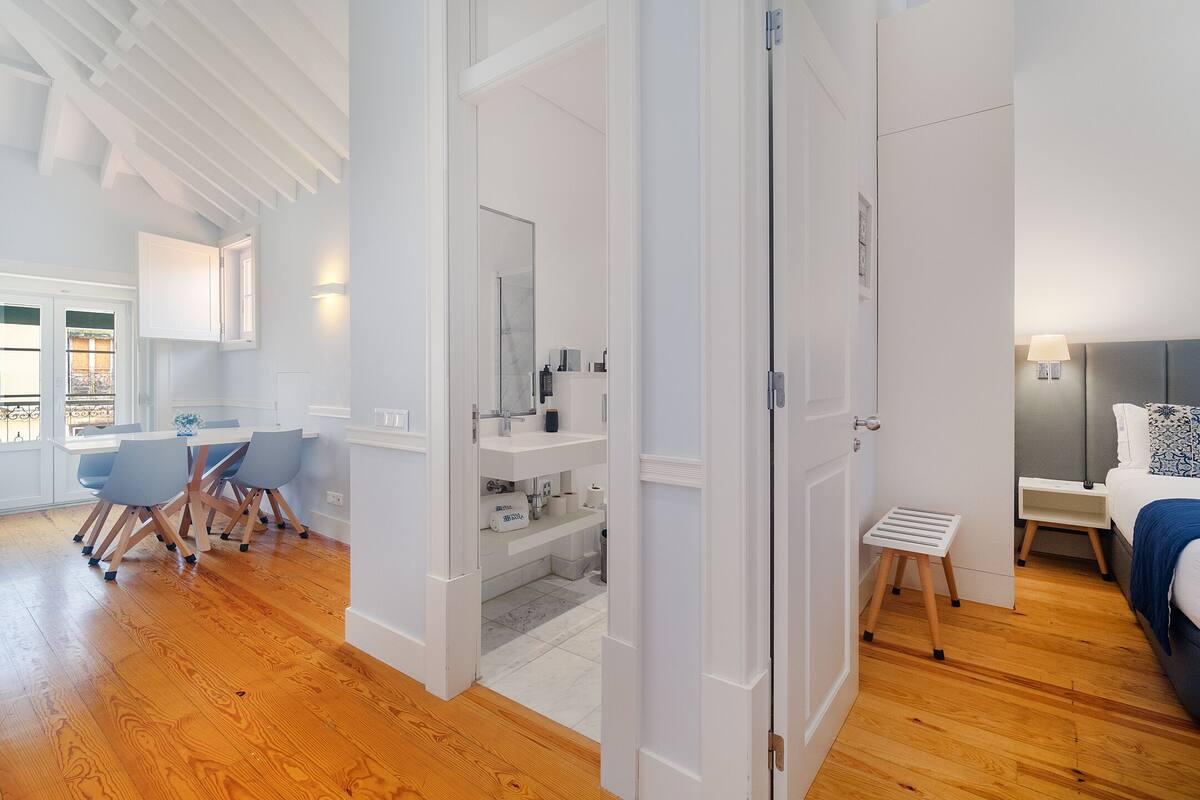 Un interior moderno con suelo de madera, un área de comedor con sillas, una cocina con armarios blancos y un dormitorio con cama y mesita de noche.