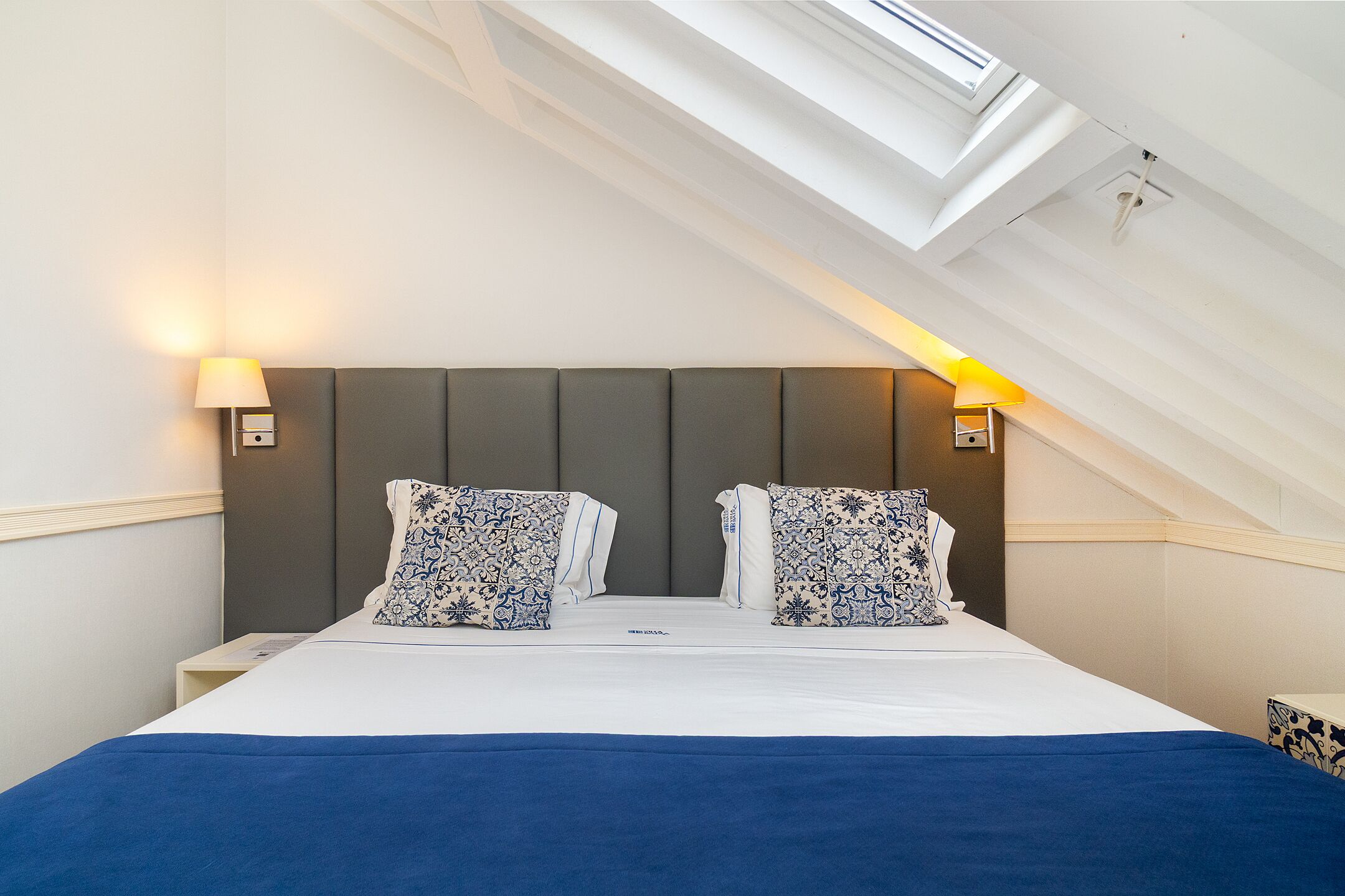 A bedroom with a skylight, a bed with blue bedding, and patterned pillows.