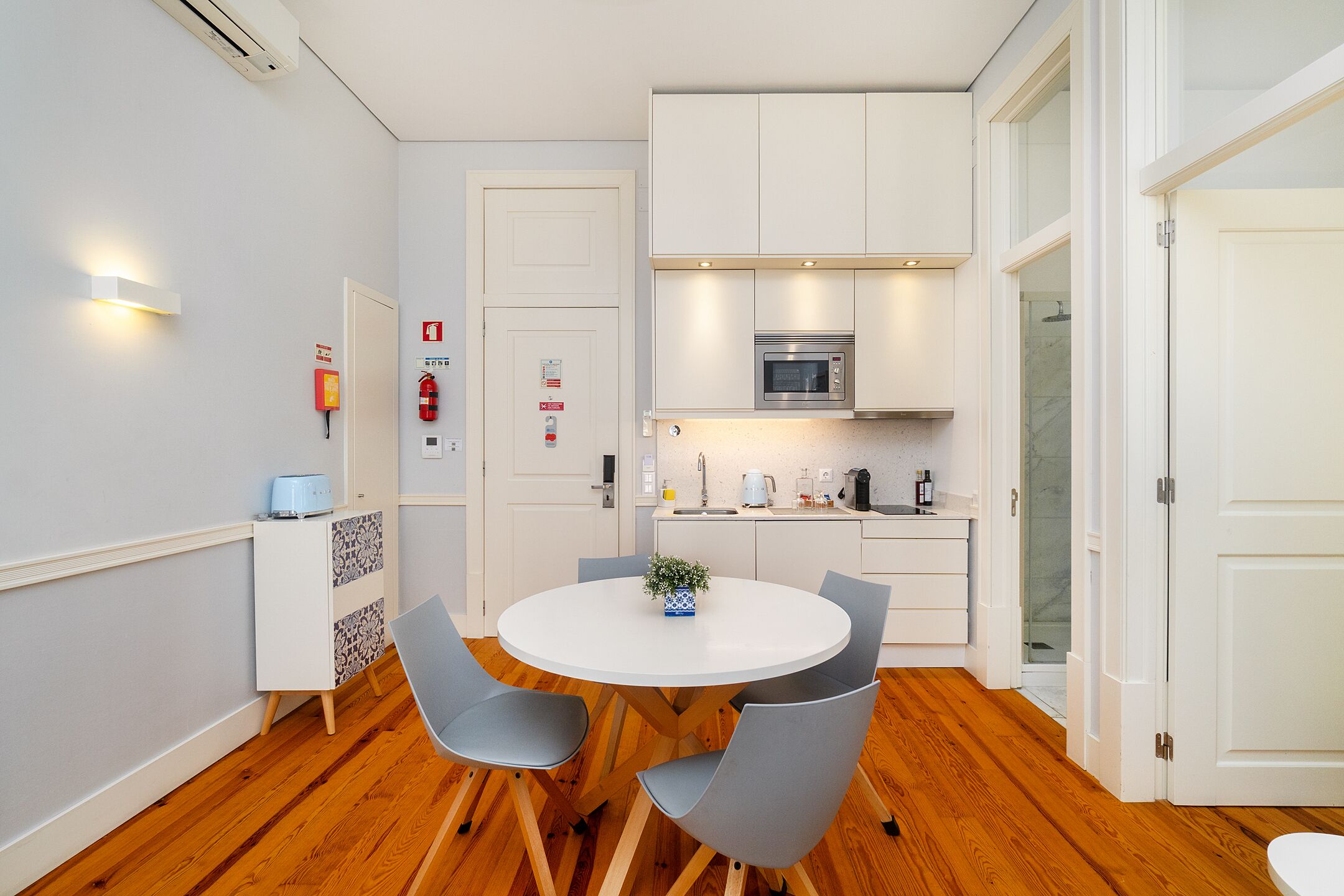 Una cocina compacta con armarios blancos, un microondas y un lavamanos. Hay una mesa de comedor con cuatro sillas y una pequeña planta en maceta.