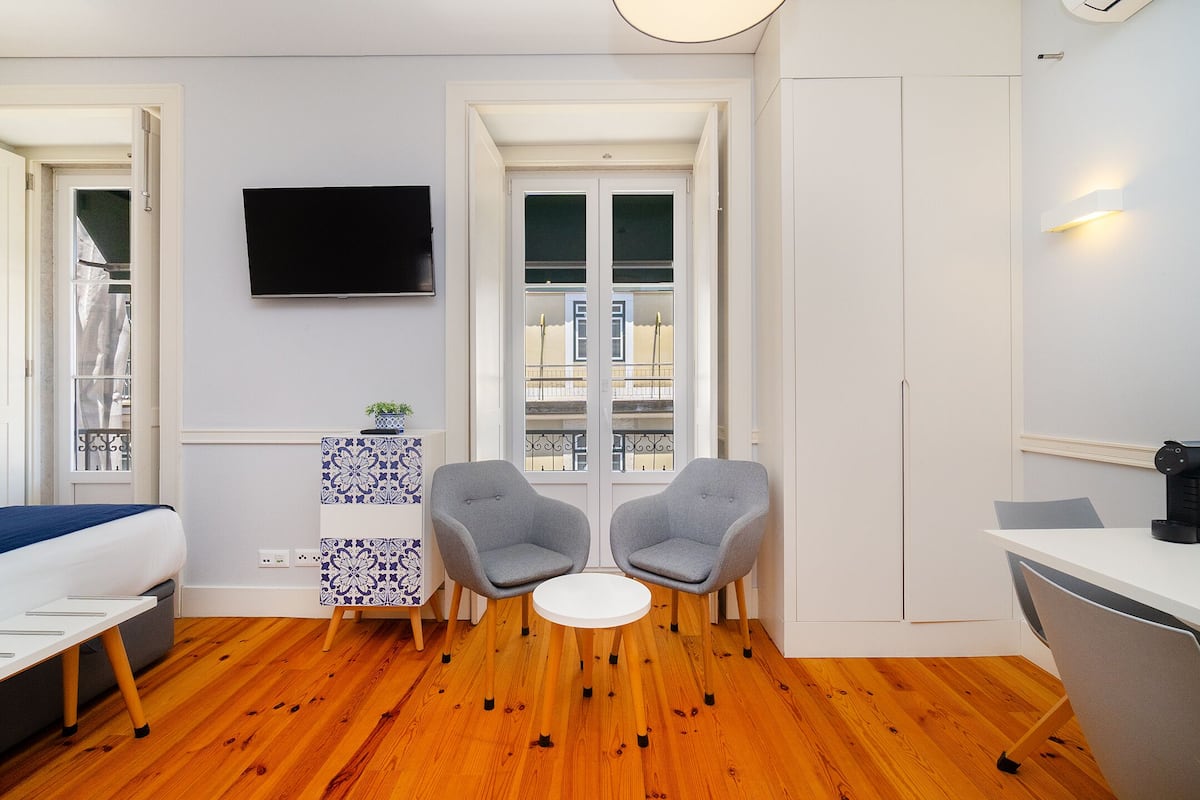 Habitación de hotel compacta con cama, una mesita pequeña con dos sillas, televisor montado en la pared y ventana con vista a edificios.
