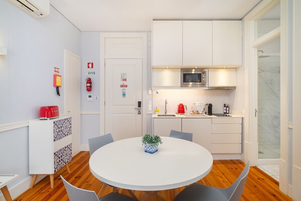 A modern kitchen with white cabinets, a microwave, and a coffee machine. There is a round dining table with chairs and a small potted plant.