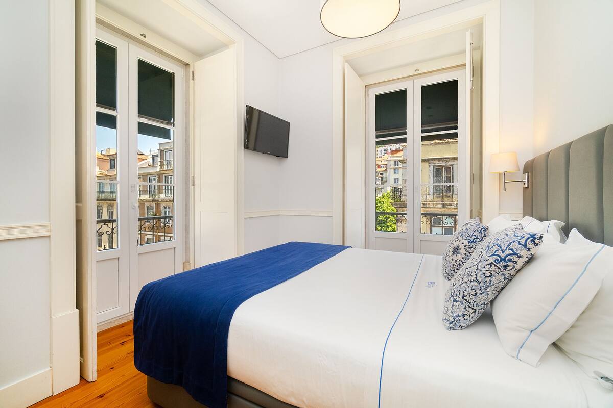 Un dormitorio con cama, televisor montado en la pared y ventana con vistas a edificios.