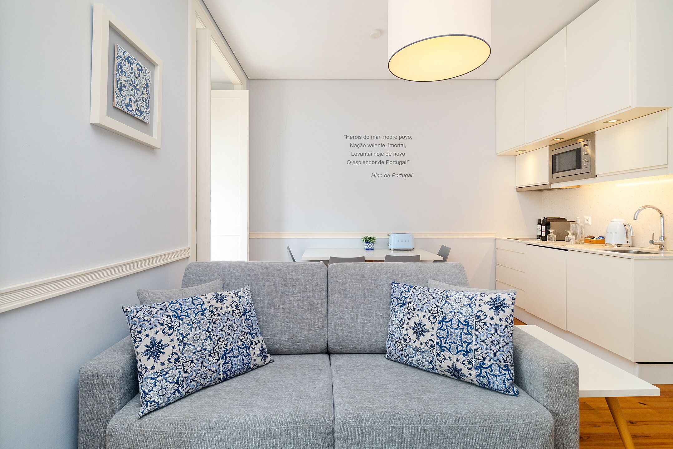 Una sala de estar moderna con un sofá gris, dos cojines estampados, una mesita con una planta y una cocina con microondas y fregadero.