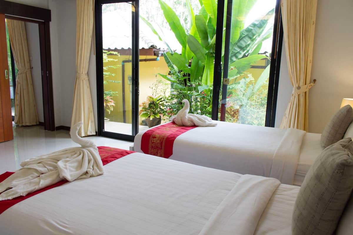 A hotel room with two beds, a red and white bedspread, a white towel arranged on one bed, and a view of a garden through a glass door.