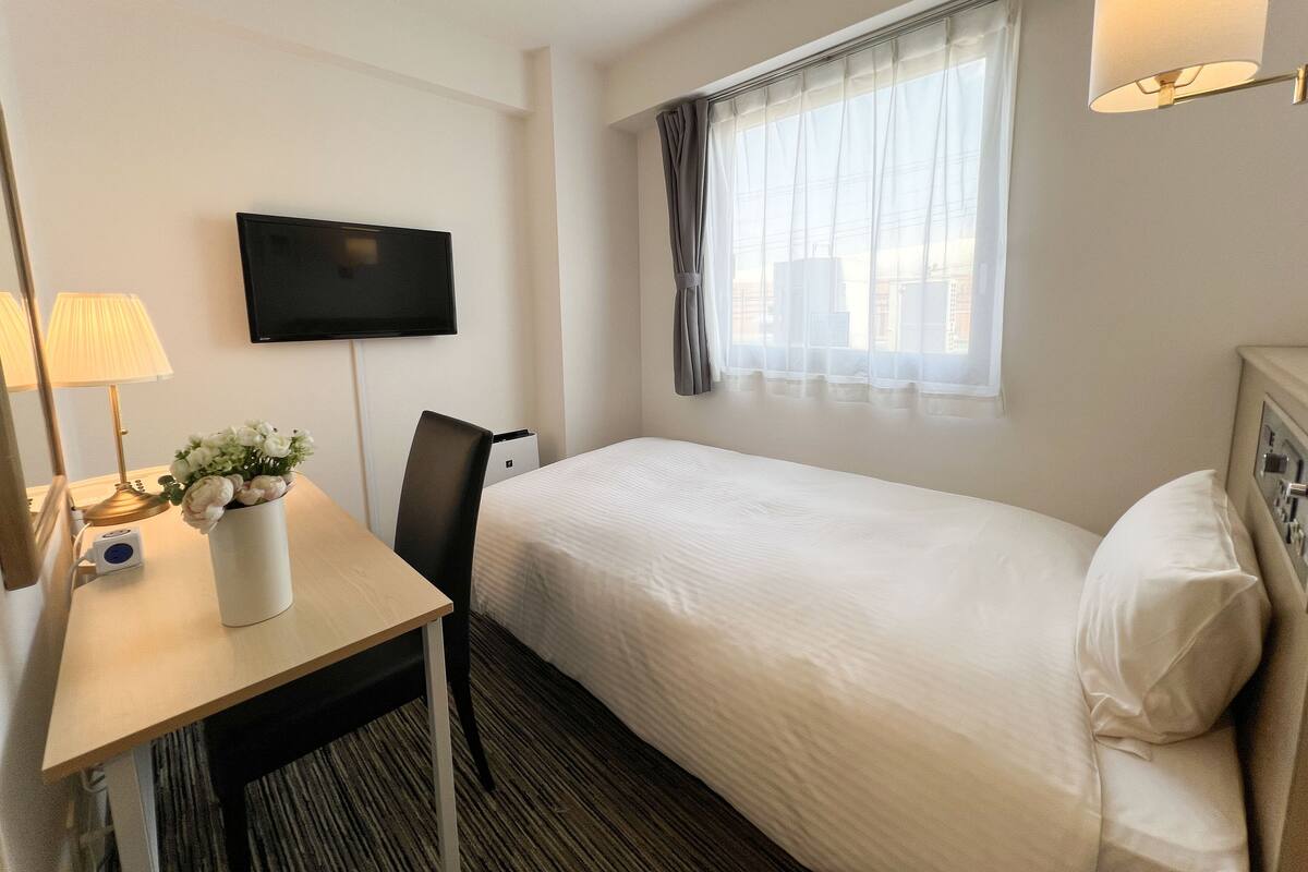 Een hotelkamer met een bed, een bureau met lamp, een aan de muur bevestigde televisie en een raam met gordijnen.