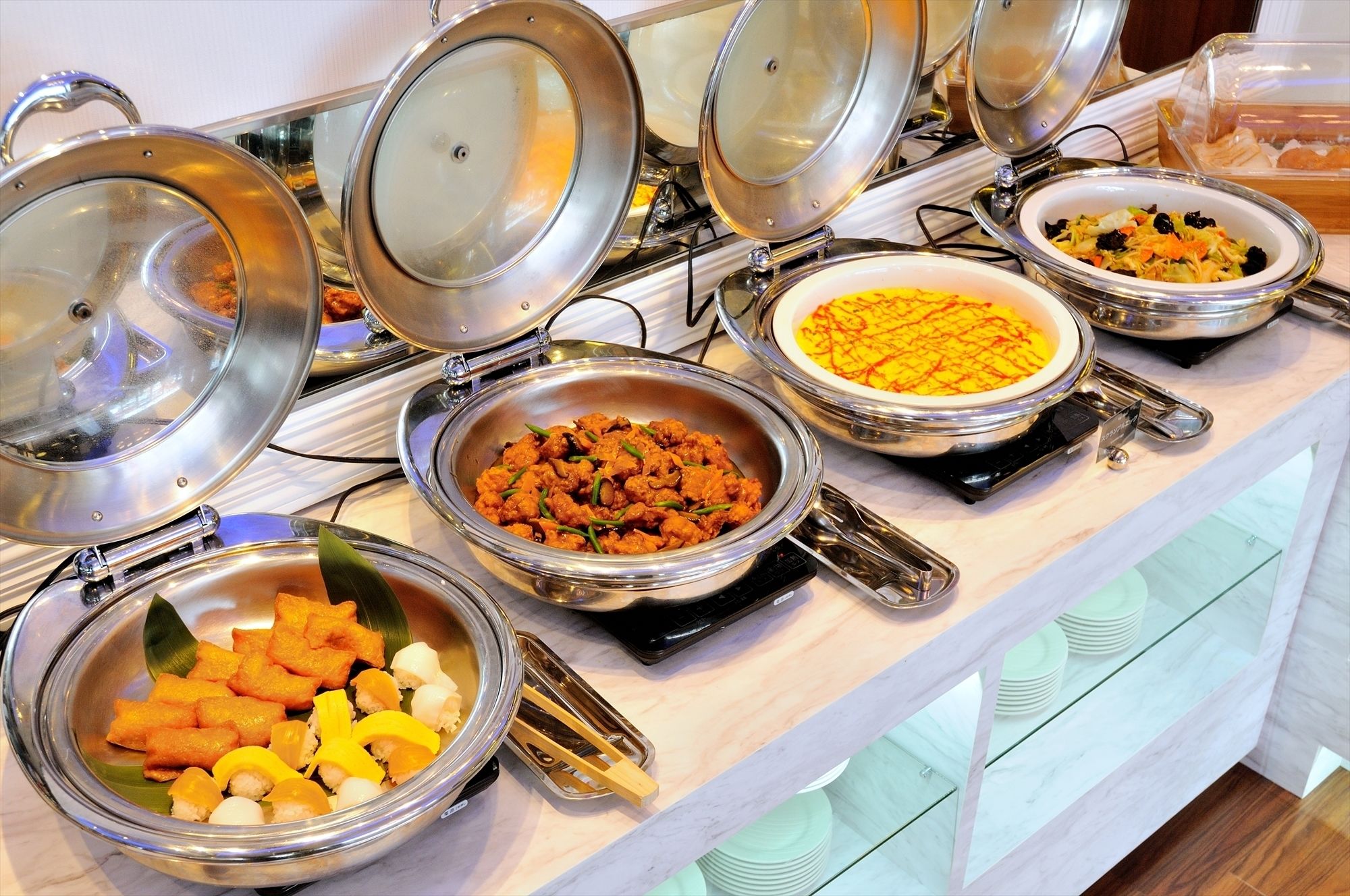 Ein Buffet mit verschiedenen Speisen in Metallschalen, einige mit Deckeln, und ein Stapel Teller auf der Theke.