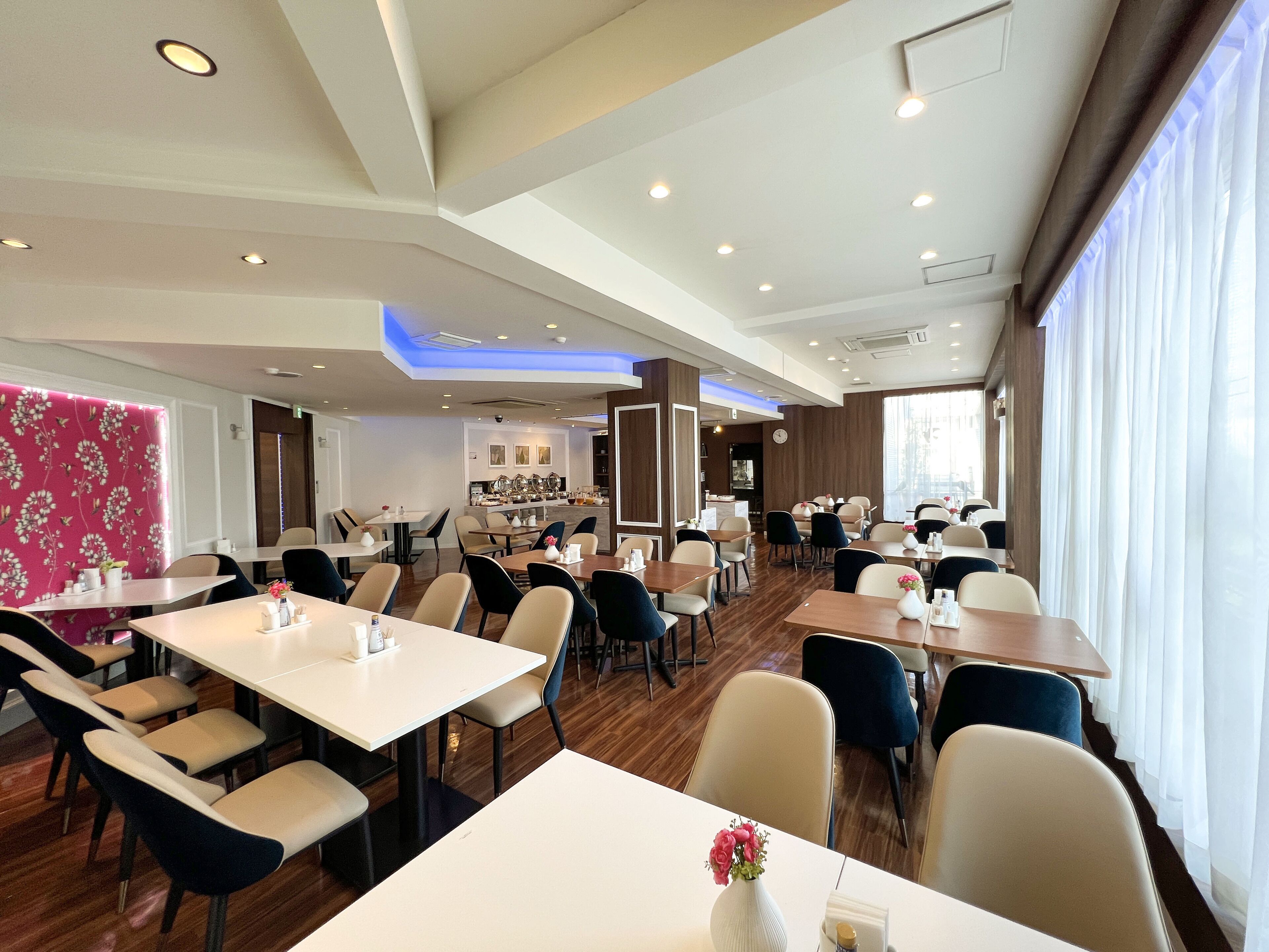 Comedor moderno de hotel con varias mesas y sillas, una cocina al fondo y ventanas grandes con cortinas.