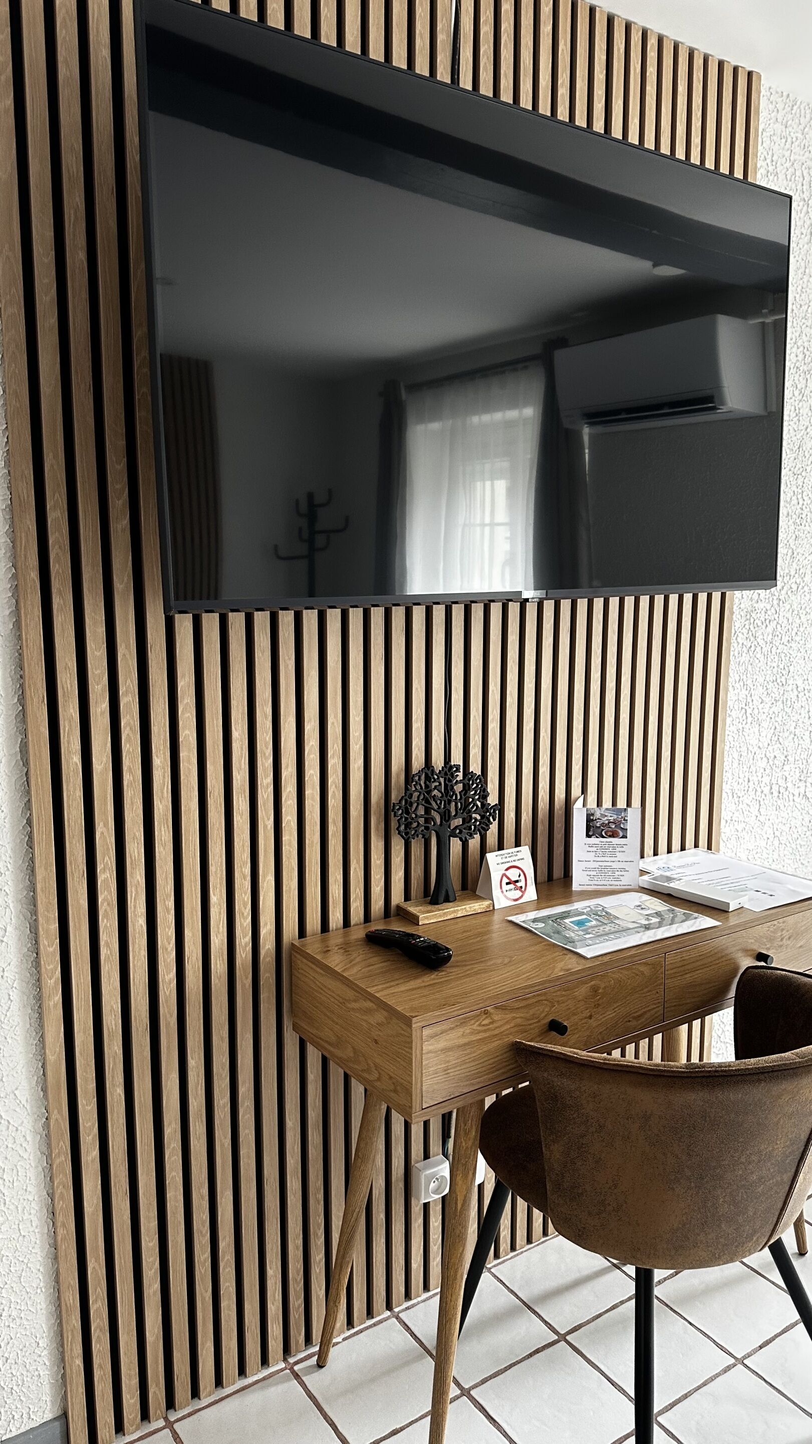 Una habitación moderna con un muro de listones de madera, un televisor montado, un escritorio con un control remoto, una silla y una pequeña mesa con un cartel.