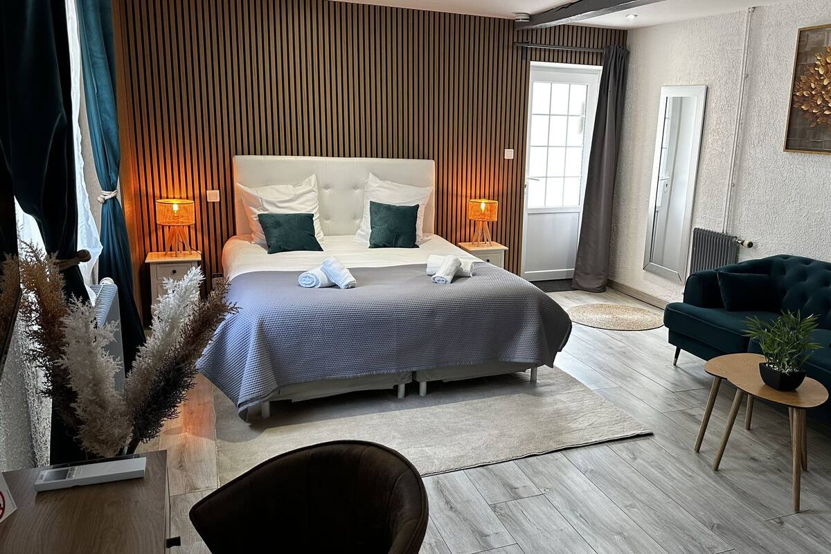 A modern hotel bedroom with a large bed, a dark green sofa, and a small round table with a plant.