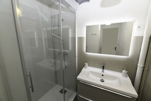Un baño moderno con cabina de ducha de vidrio, un lavamanos blanco con un espejo encima y azulejos color beige.