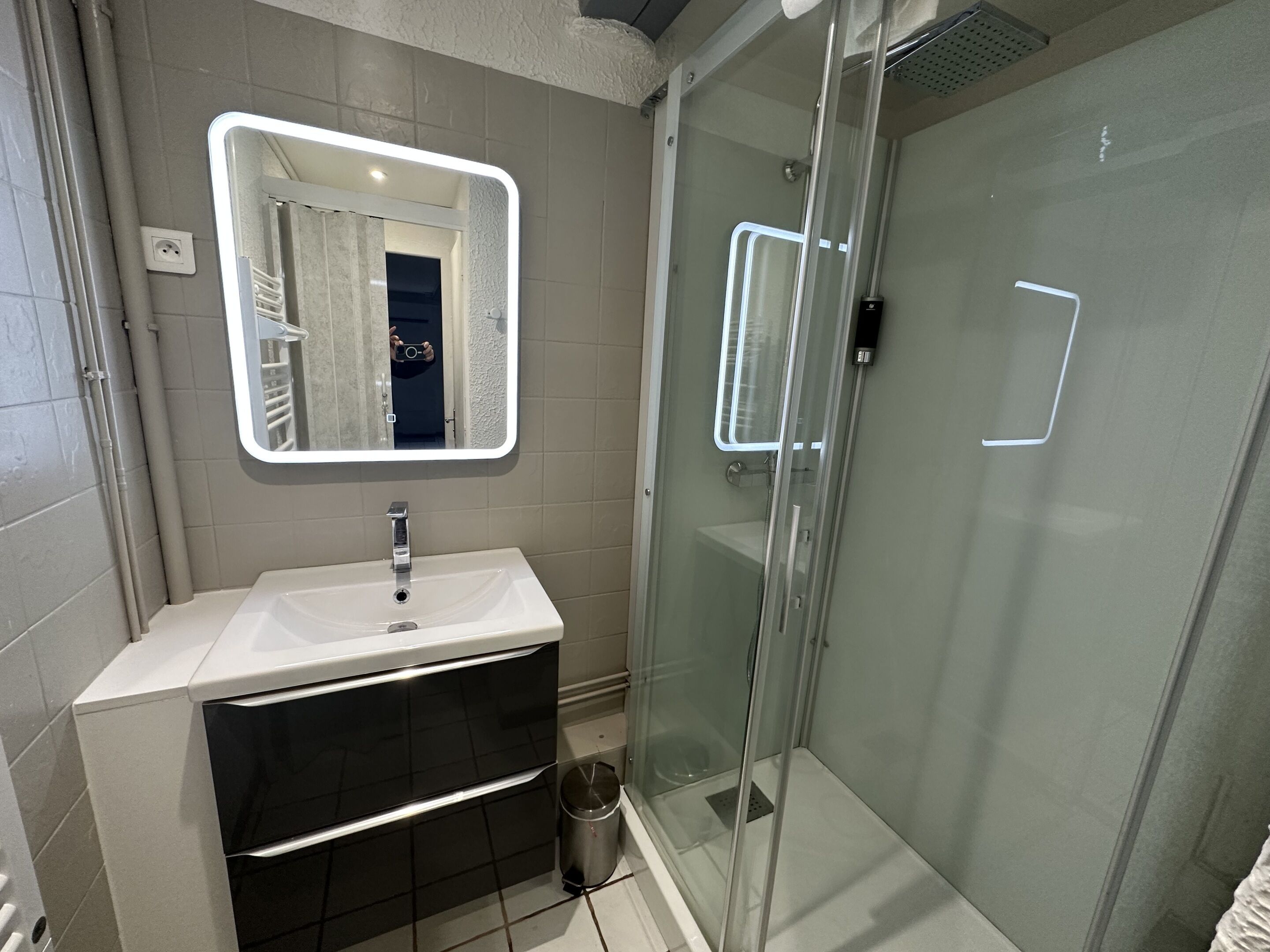 Une salle de bain moderne avec une douche à parois de verre, un lavabo blanc avec un miroir au-dessus et une poubelle.
