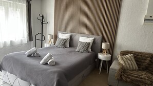 Uma cama bem arrumada com um espalhador cinzento e duas almofadas, uma luminária de cabeceira, um cabideiro e um cobertor macio numa cadeira.