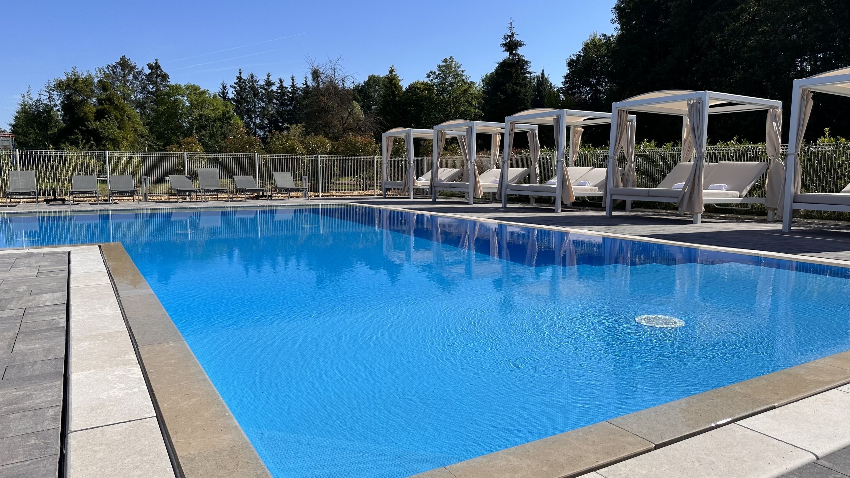 A clear blue pool with lounge chairs and canopies, surrounded by a metal fence and trees.