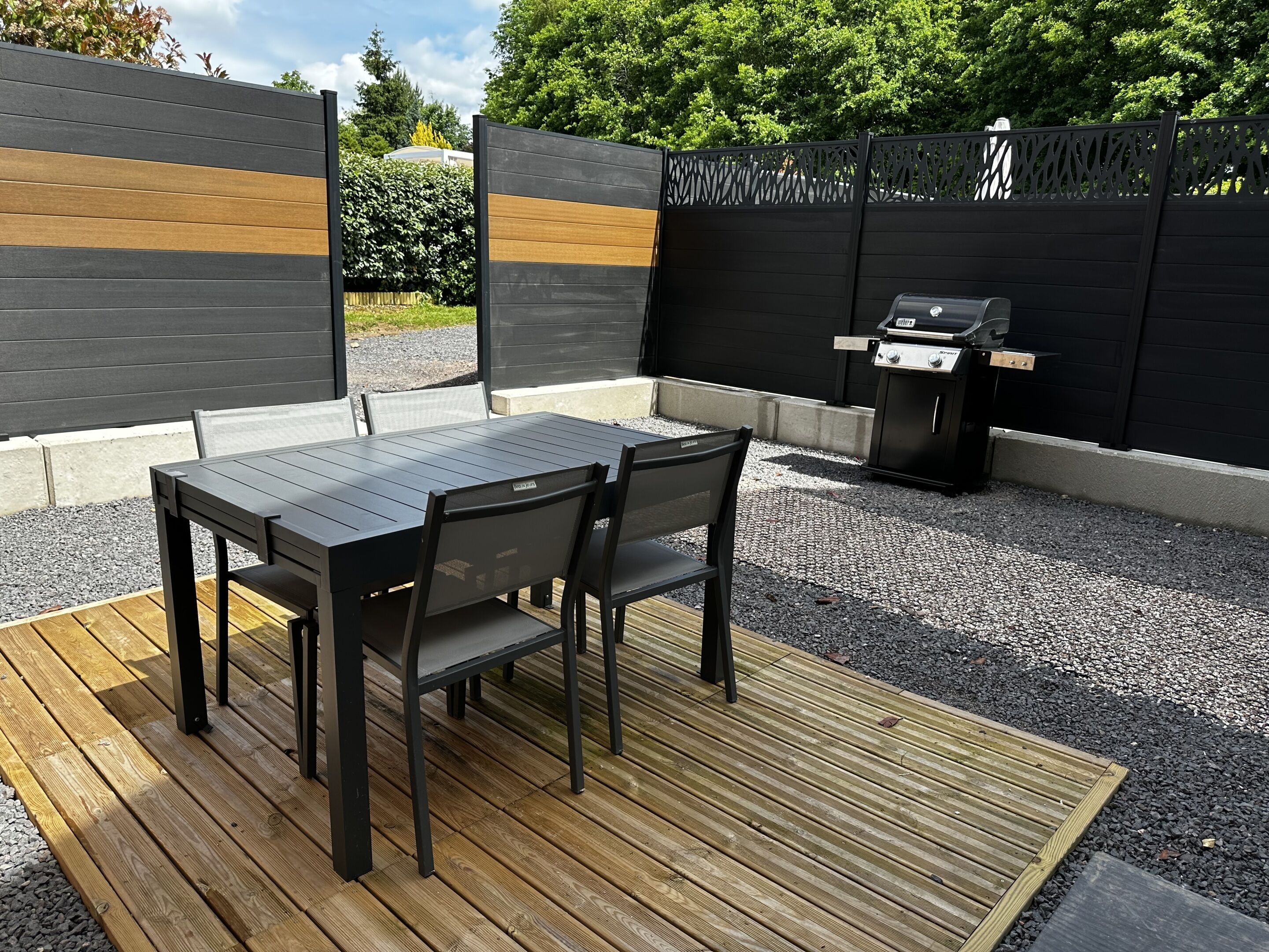 Un patio al aire libre con mesa y sillas de madera, una parrilla y un cerco decorativo.
