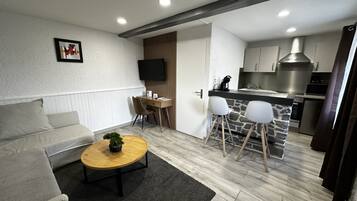 Un espacio habitable compacto con un sofá, una mesa de centro con una planta, un televisor montado en la pared, una zona de cocina con estufa y armarios, y un comedor con sillas.
