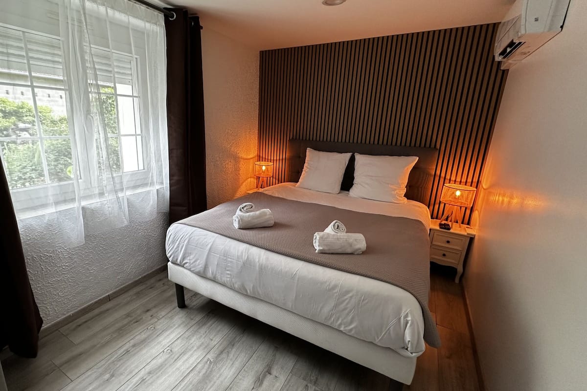 A modern hotel room with a large bed, two bedside tables with lamps, a window with sheer curtains, and a view of greenery outside.