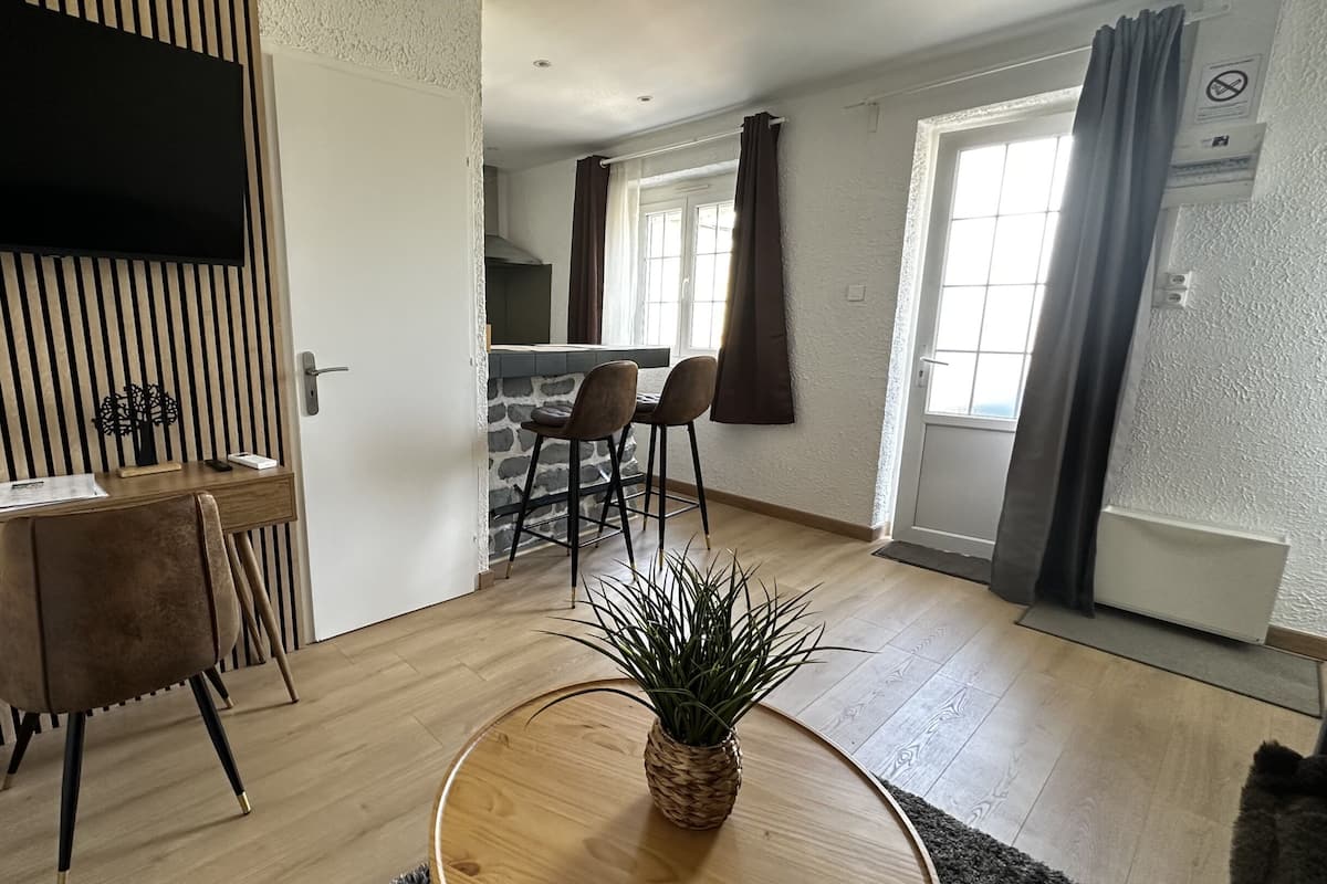 A modern living room with a wooden dining table, two chairs, a TV mounted on a slatted wall, a kitchen area with a window, and a potted plant on a round wooden table.
