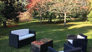 An outdoor seating area with a sofa and chairs, a fire pit, and a grassy lawn with trees in the background.