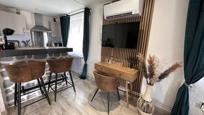 Uma cozinha moderna com ilha de pedra, banquetas, uma escrivaninha de madeira e televisão na parede.