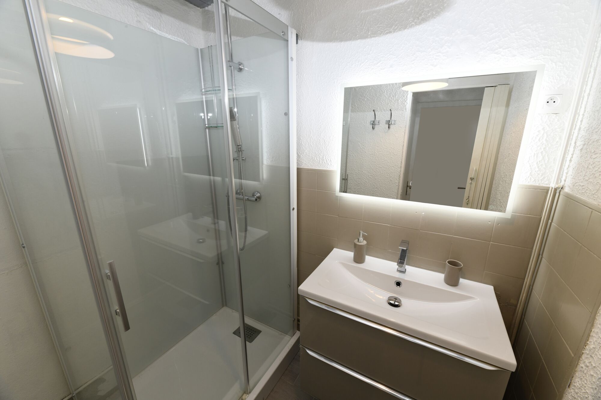 Un baño con ducha de vidrio, un lavamanos con espejo y azulejos color crema.