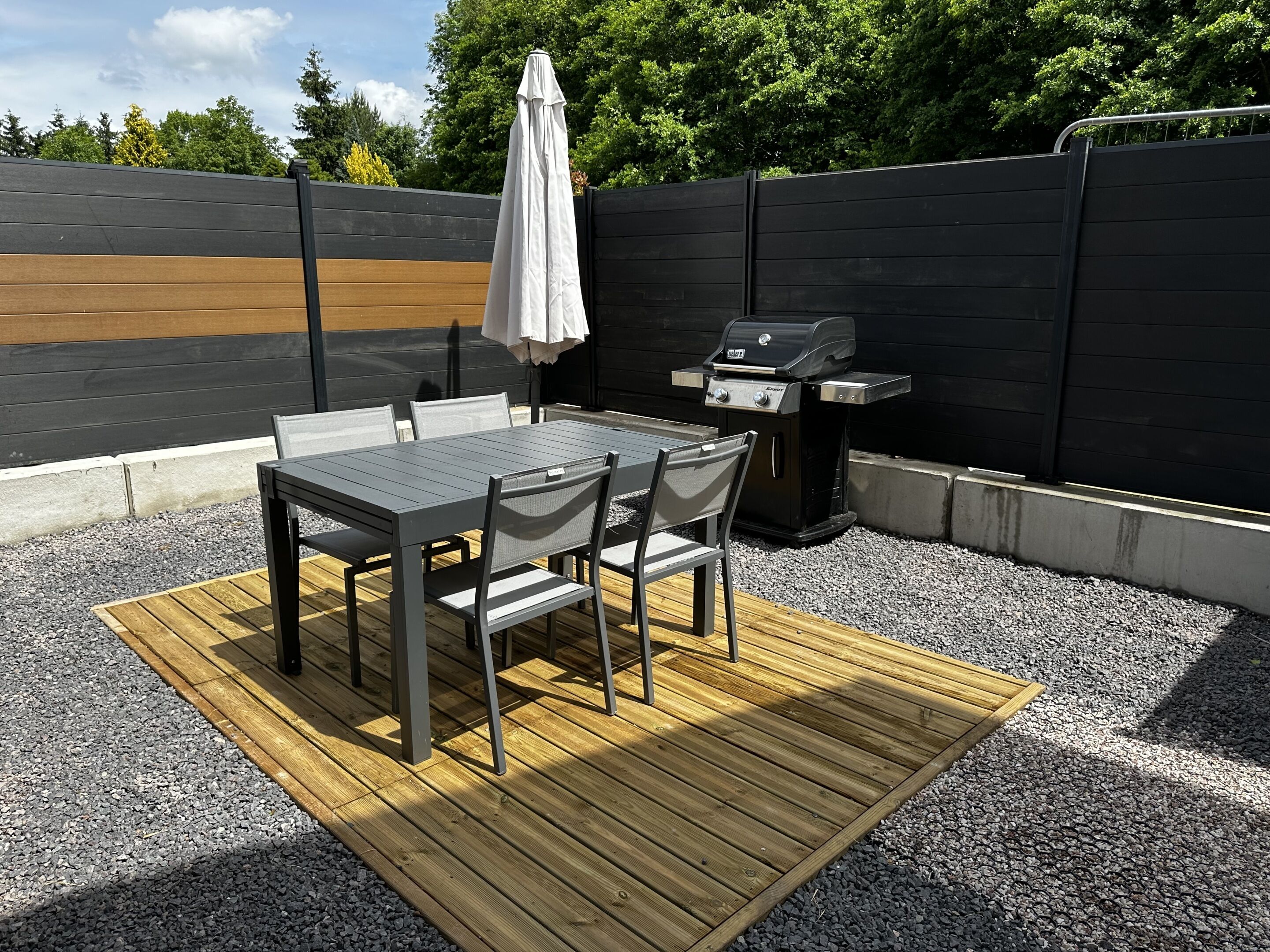 An outdoor patio with a table and chairs, a grill, and a wooden deck.