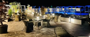 Un patio al aire libre con mobiliario de mimbre, plantas en macetas y una zona de piscina con iluminación azul.