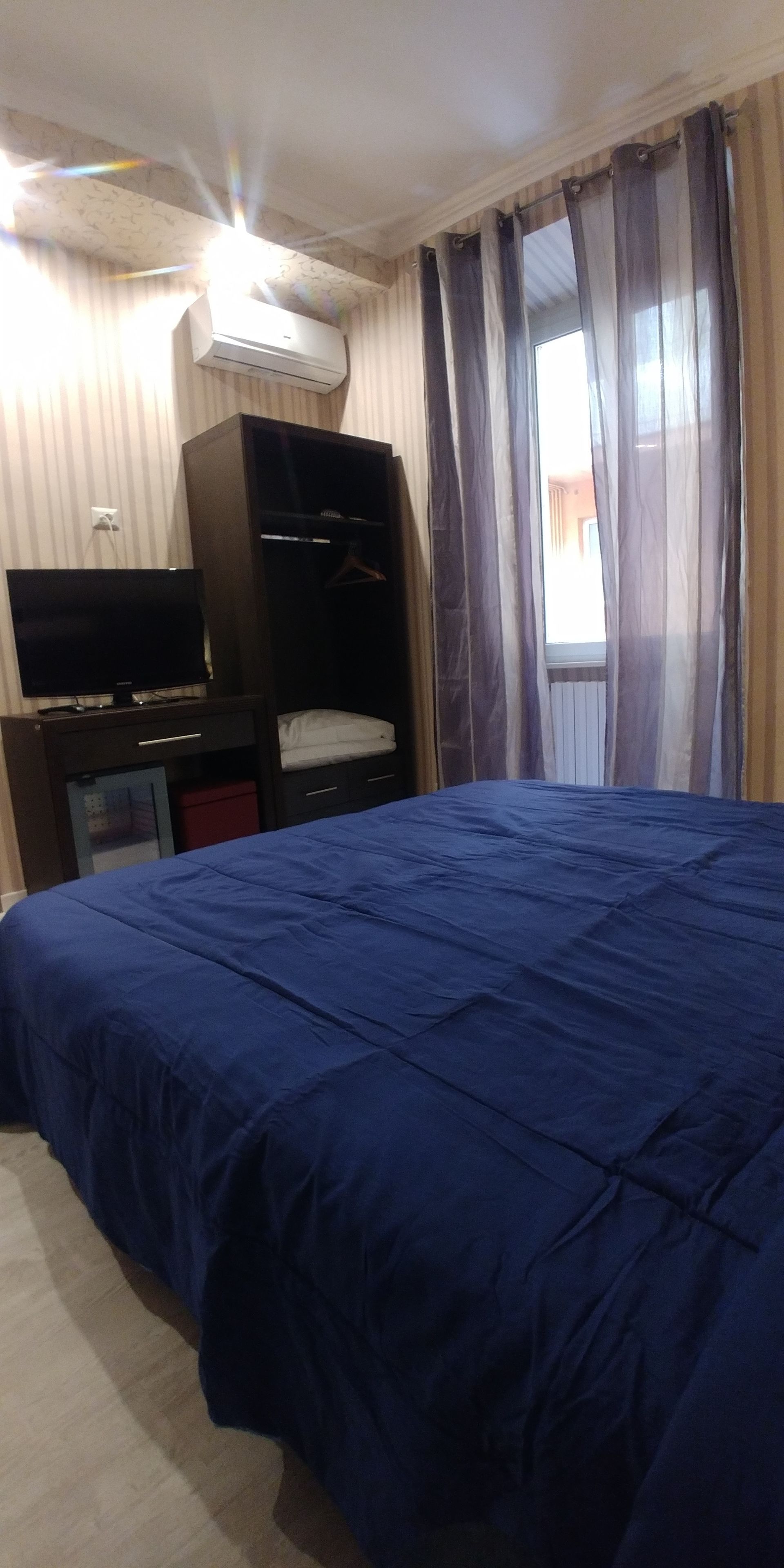 A bedroom with a large bed, a TV on a dresser, an air conditioner, and a window with curtains.
