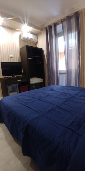 Een slaapkamer met een groot bed, een televisie op een dressoir, een airconditioner en een raam met gordijnen.
