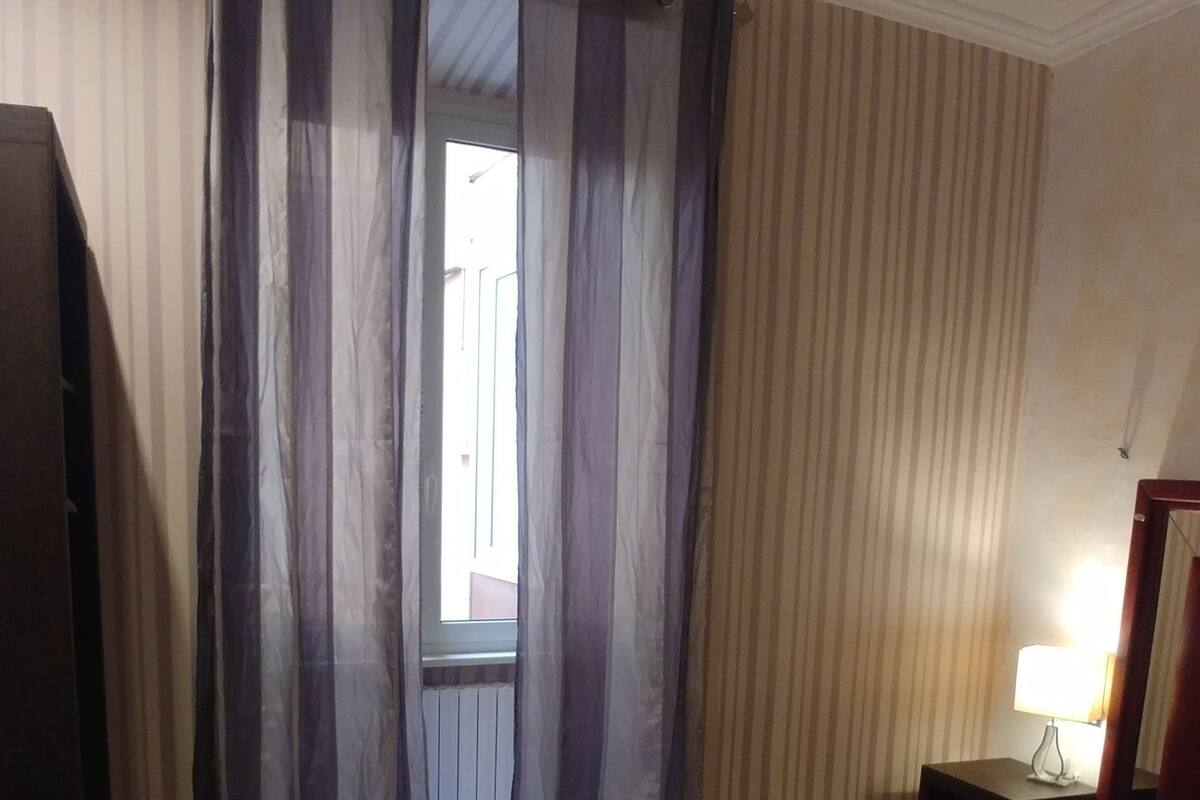 Una habitación de hotel con cama, cortinas, mesita de noche con lámpara y cómoda de madera.
