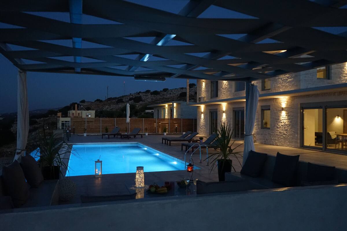 A modern outdoor pool area with a pergola, lounge chairs, and a view of a hillside.
