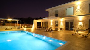 A modern outdoor pool area with a stone facade, lounge chairs, and a covered seating area. - Eva Villas (Rethymno)