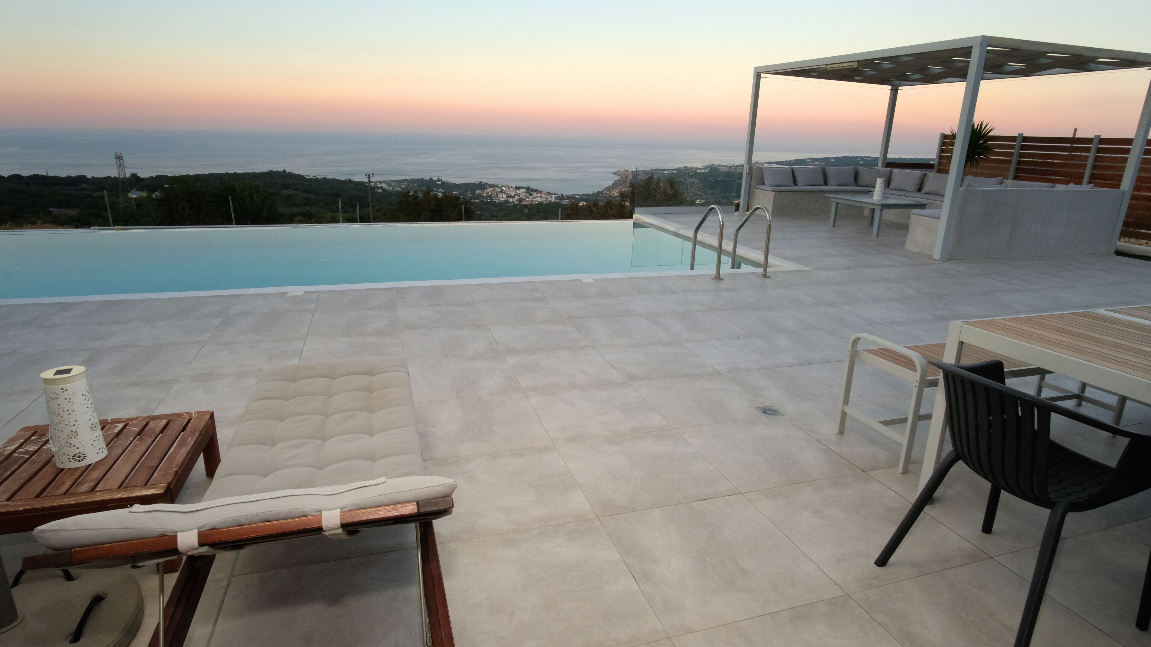 Zwembadgedeelte met uitzicht op zee bij zonsondergang, buitentafels en een pergola.