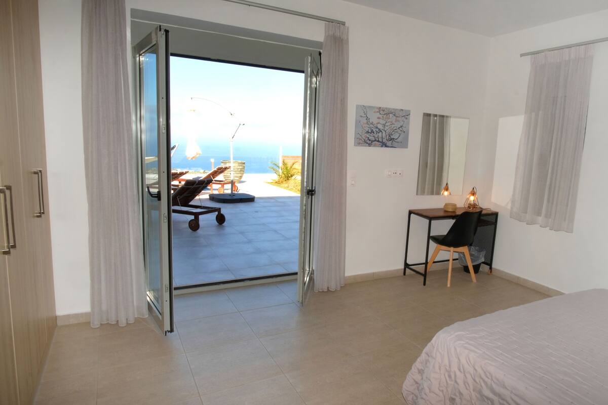 A hotel room with a bed, a desk with a chair, a window with curtains, and a view of the ocean.