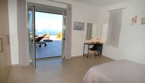 A bedroom with a bed, a desk with a chair, a window with curtains, and a view of the outdoors. - Eva Villas (Rethymno)