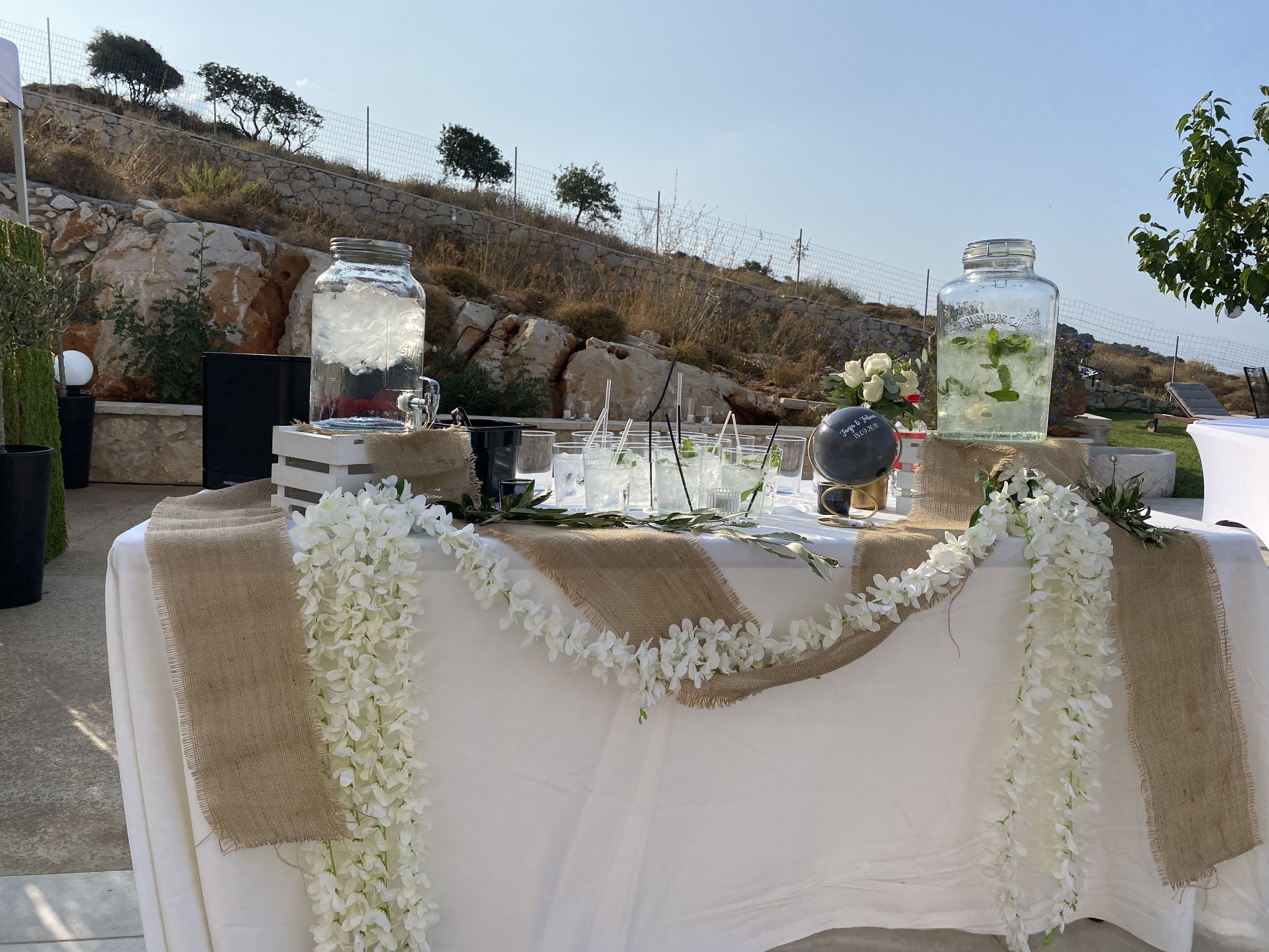 An outdoor event setup with a table covered in white cloth and burlap, adorned with white flowers.