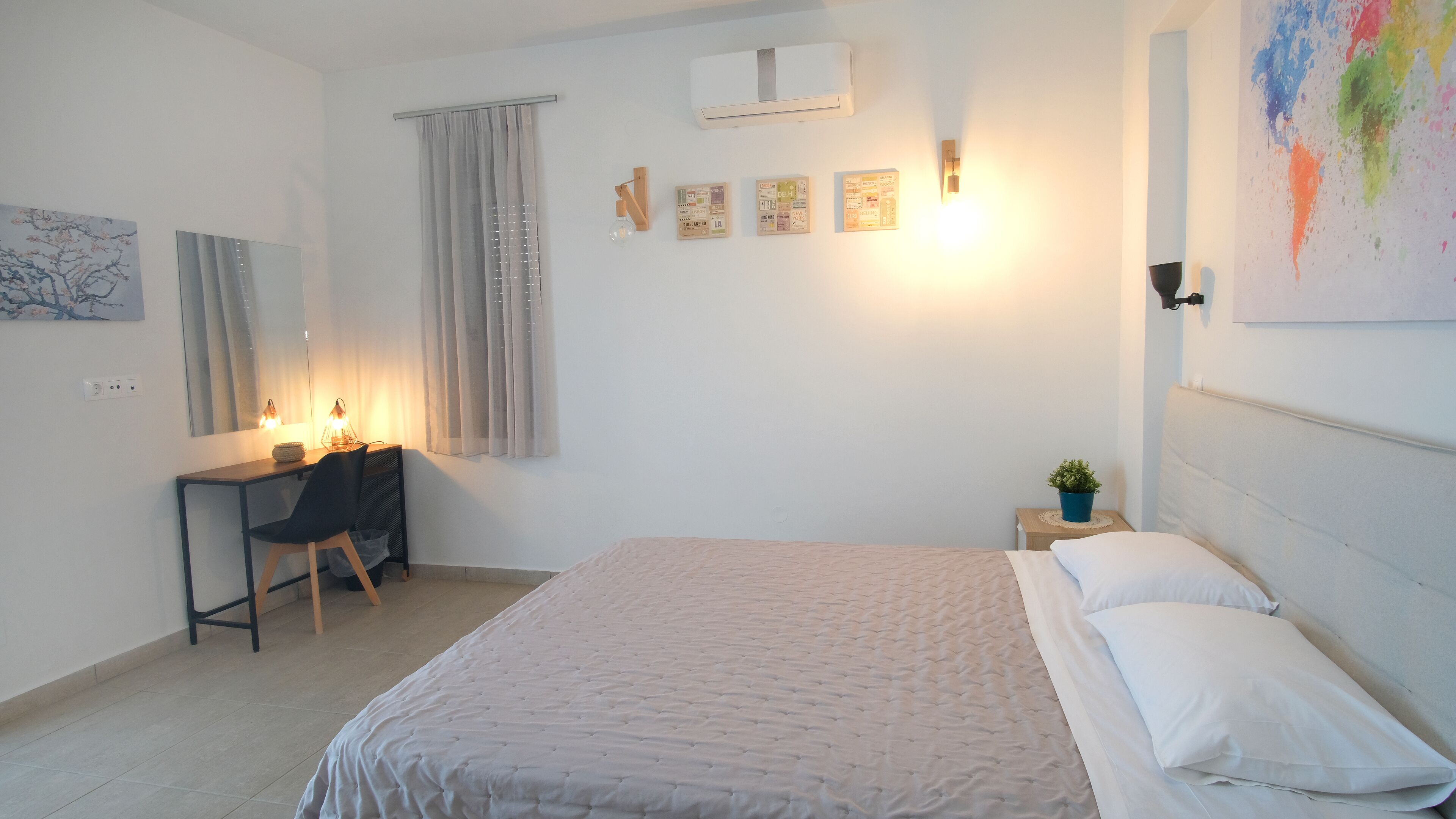 Un dormitorio con cama, escritorio y silla. Se observa una ventana con cortinas, un equipo de aire acondicionado y una lámpara en la pared.